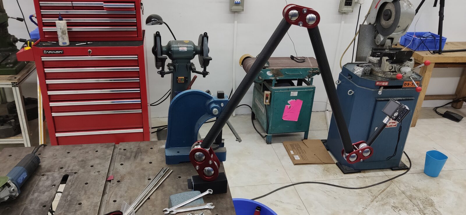 Long arm with red joints reaching across a workshop floor with a red tool chest, grinder and machine tools behind it.