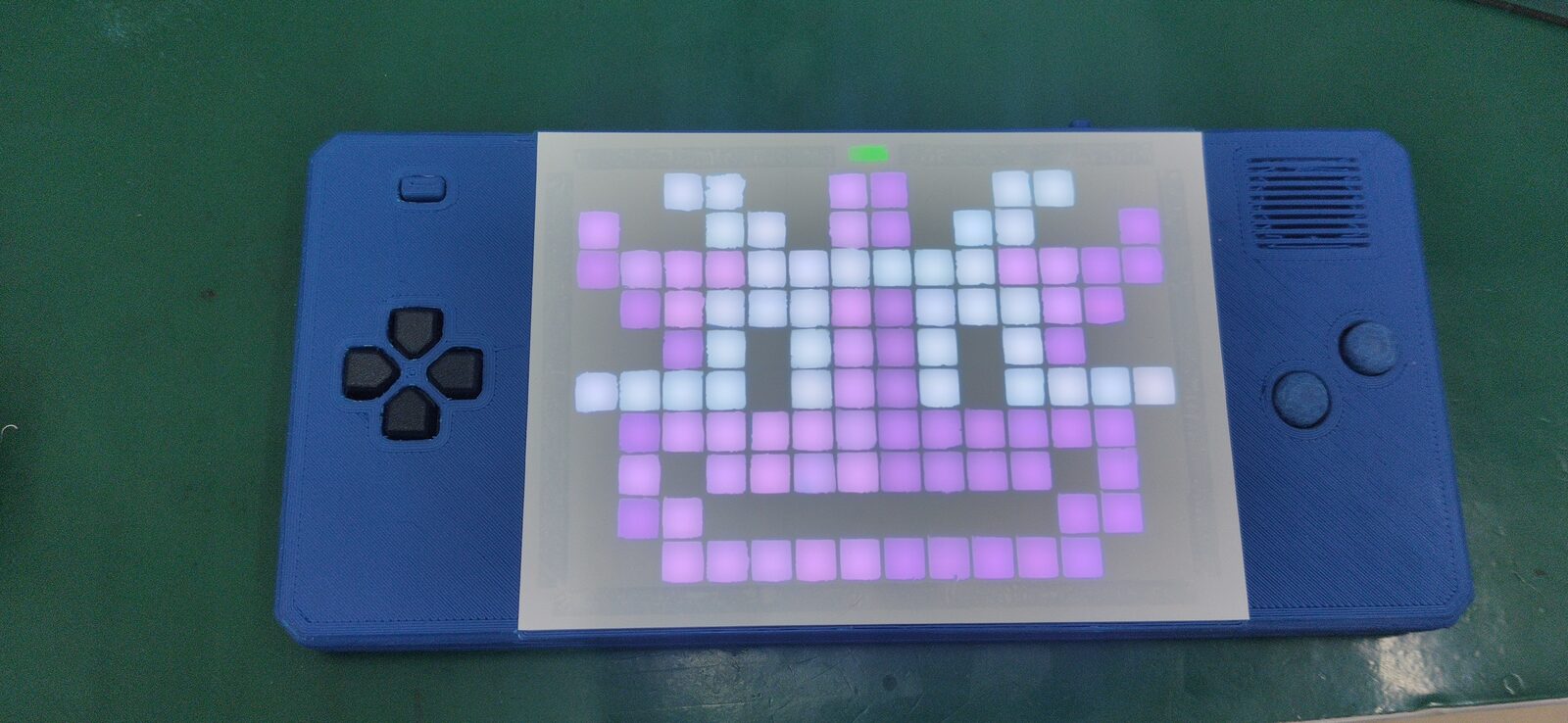 Blue handheld on a green mat with a grid display lit in purple showing a smiling pixel face.